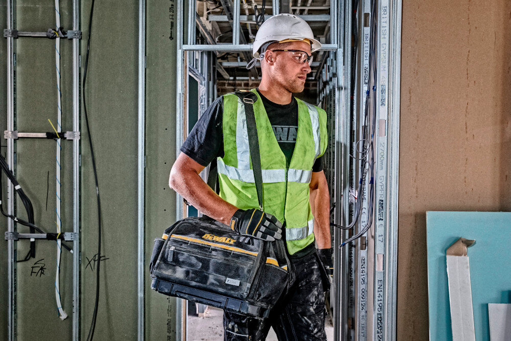 Torba narzędziowa DEWALT DWST82991-1 plac budowy osoba niesie torbę zawieszoną na ramię w tle rusztowania elementy konstrukcyjne praktyczne przechowywanie uniwersalne zastosowanie komfort transportu