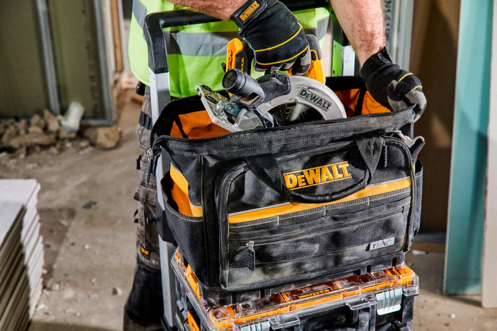 Torba narzędziowa DEWALT DWST82991-1 plac budowy osoba wyciąga z wnętrza torby elektronarzędzie w tle gruz płyt kartonowo-gipsowe z mocnej tkaniny poliestrowej o gęstości 1200 denier duża odporność na uszkodzenia mechaniczne podwójne szwy stalowe nity długotrwałe użytkowanie