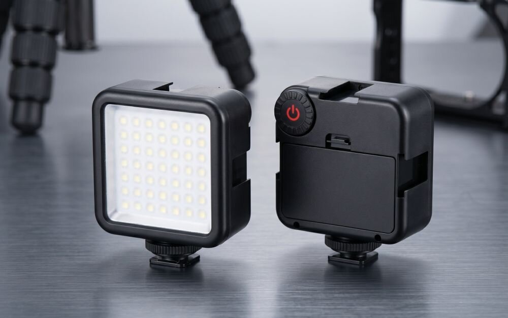 Na obrazie widoczne są dwie czarne kompaktowe lampy LED, jedna z przodu z widocznymi diodami, druga z tyłu z przyciskiem zasilania. Obie lampy znajdują się na metalowej powierzchni.