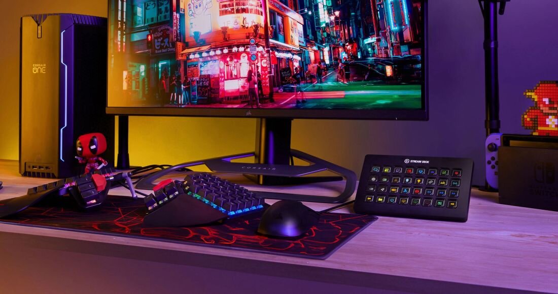 Na biurku znajduje się komputer z dużym monitorem, klawiaturą, myszką i panelem z przyciskami oznaczonym logo \'Stream Deck\'. W tle widoczna jest kolorowa scena z gry lub filmu na ekranie monitora.