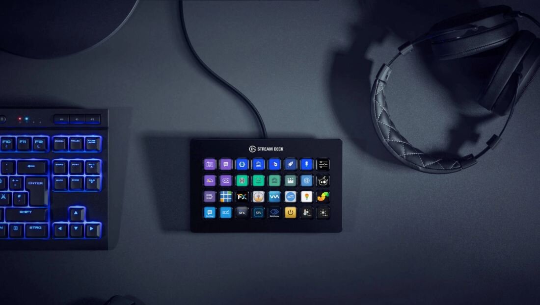 Na ciemnym biurku znajduje się panel z wieloma podświetlanymi przyciskami, na którym widnieje napis \'STREAM DECK\'. Obok panelu leży klawiatura z niebieskim podświetleniem oraz słuchawki.