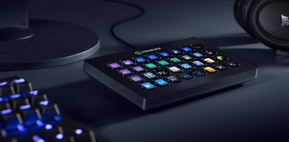 Na obrazie widoczny jest panel z wieloma podświetlanymi przyciskami, na którym znajduje się napis \'STREAM DECK\'. W tle widać fragmenty klawiatury i słuchawek.