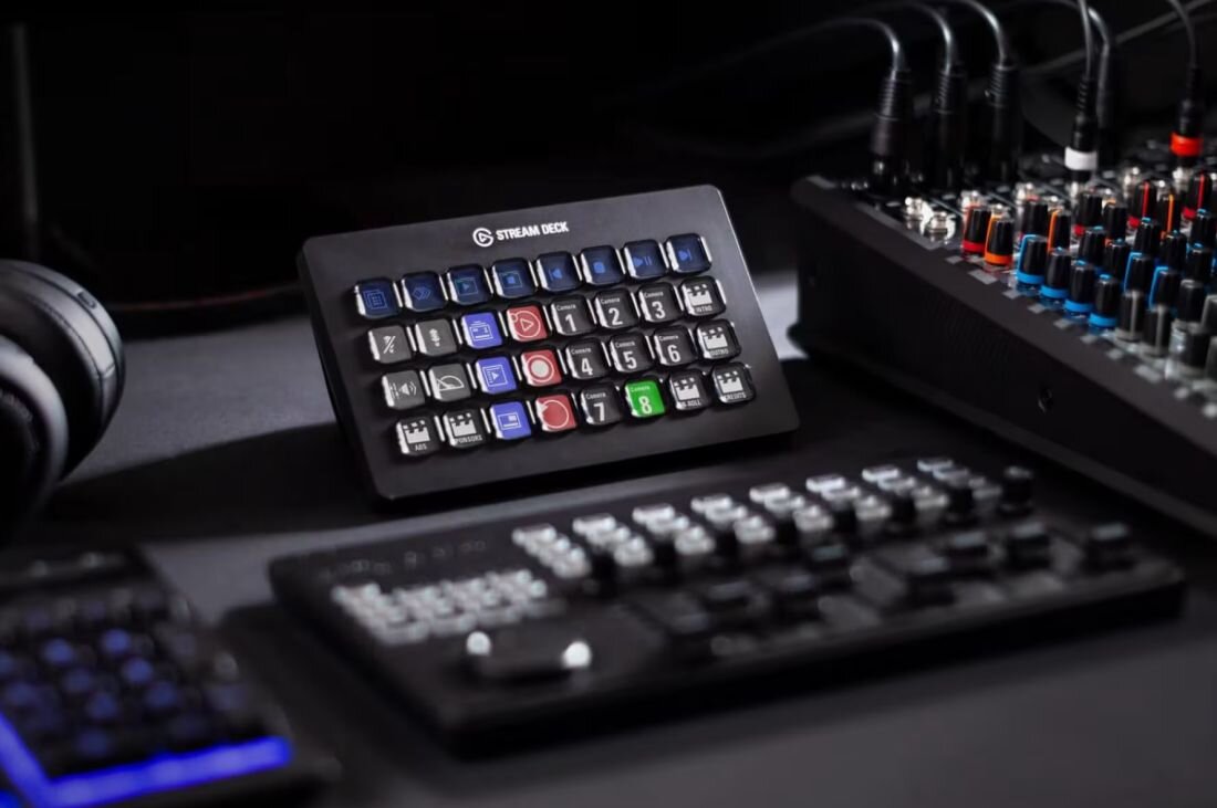 Na obrazie widoczny jest panel z przyciskami oznaczonymi różnymi ikonami i numerami, z napisem \'STREAM DECK\' na górze. W tle znajdują się słuchawki i sprzęt audio z pokrętłami i kablami.