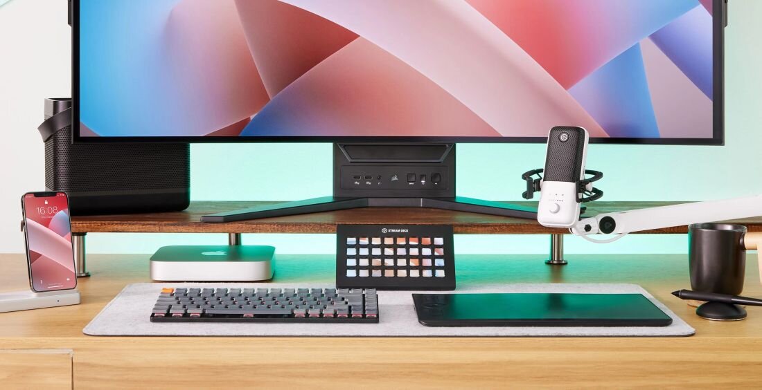Na biurku znajduje się monitor, klawiatura, panel z przyciskami, mikrofon oraz smartfon. Panel ma logo \'ELGATO\' i jest umieszczony przed monitorem.