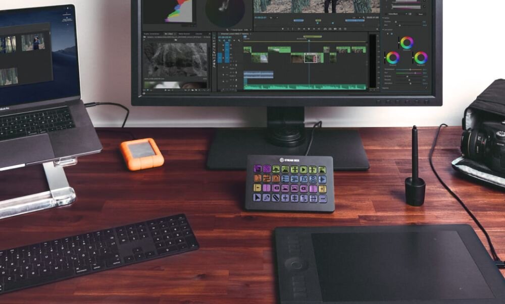 Na biurku znajduje się panel z wieloma przyciskami, monitor z wyświetlonym oprogramowaniem do edycji wideo, laptop, klawiatura, tablet graficzny i aparat fotograficzny. Na panelu widoczne jest logo \'Elgato\'.