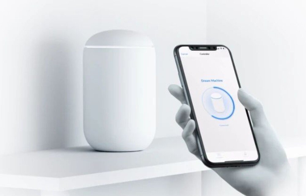 Router UBIQUITI UDM-EU Dłoń trzymająca smartfon wyświetlający ekran konfiguracji urządzenia, podczas gdy w tle na białej półce stoi właściwy sprzęt w kształcie kapsuły