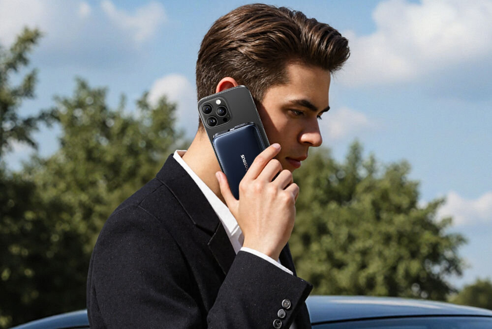 Powerbank BASEUS PicoGo AM61 QI2.2 10000 mAh 45W Czarny Mężczyzna w marynarce stoi na zewnątrz w jasny dzień i rozmawia przez telefon. Jego twarz jest zamazana, a w ręce trzyma smartfon z zamocowanym na nim powerbankiem marki Baseus.

