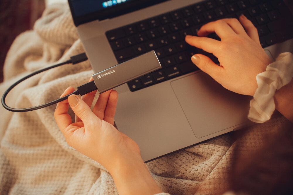 Osoba trzyma szary dysk z logo \'Verbatim\', podłączony do laptopa. Ręce osoby są na klawiaturze laptopa, a tło stanowi jasny koc.