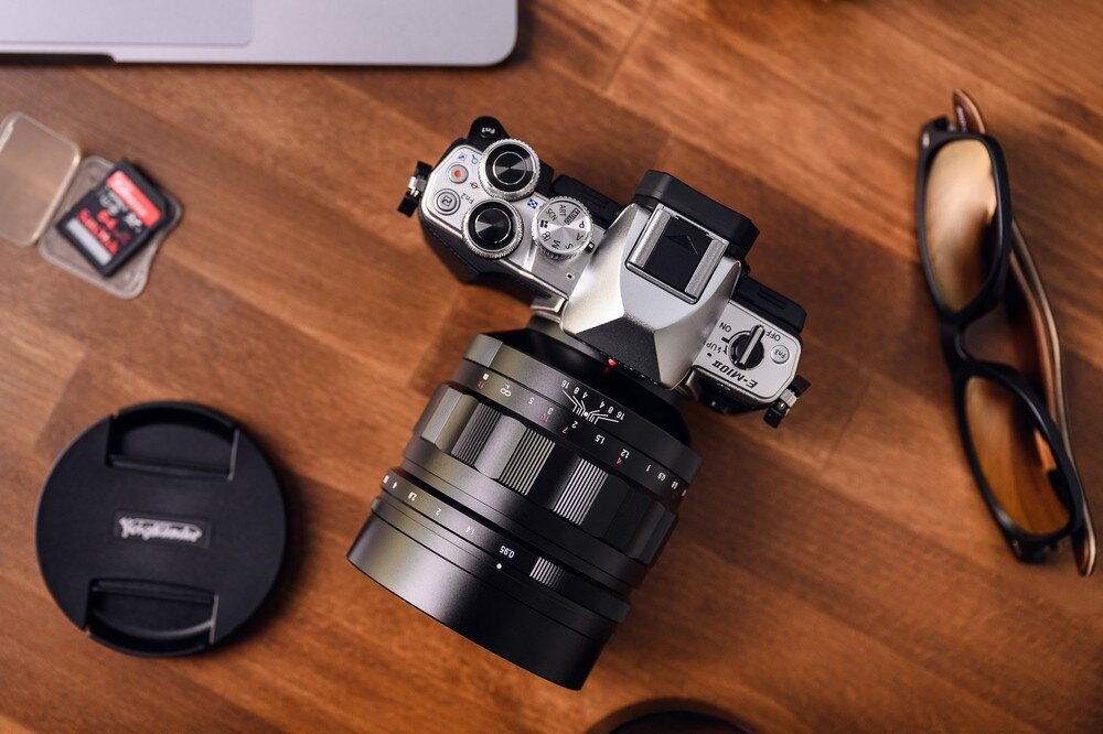 Obiektyw VOIGTLANDER 60 mm f-0.95 Nokton regulacja przysłony f/0,95–f/16 Aparat Micro 4/3 z zamontowanym obiektywem Voigtländer Nokton 60 mm f/0,95, leżący na biurku obok akcesoriów takich jak karta pamięci i osłona obiektywu, podkreślające solidność konstrukcji i elegancki design w otoczeniu fotograficznym.