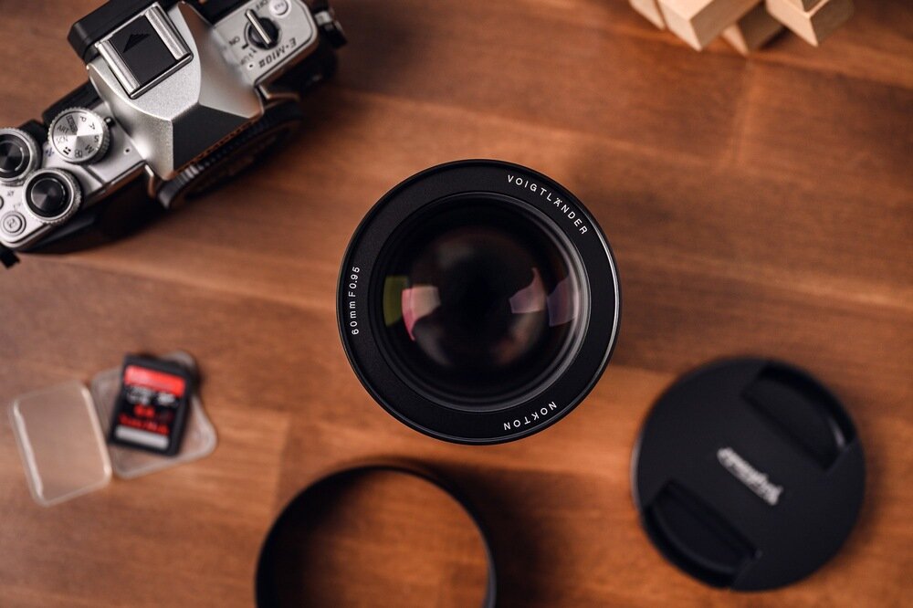 Obiektyw Voigtländer Nokton 60 mm f/0,95 Micro 4/3 Kompatybilność widziany z góry, z detalami soczewek i wyraźnym oznaczeniem parametrów, ułożony na drewnianym biurku w otoczeniu aparatu, karty pamięci i dekielka