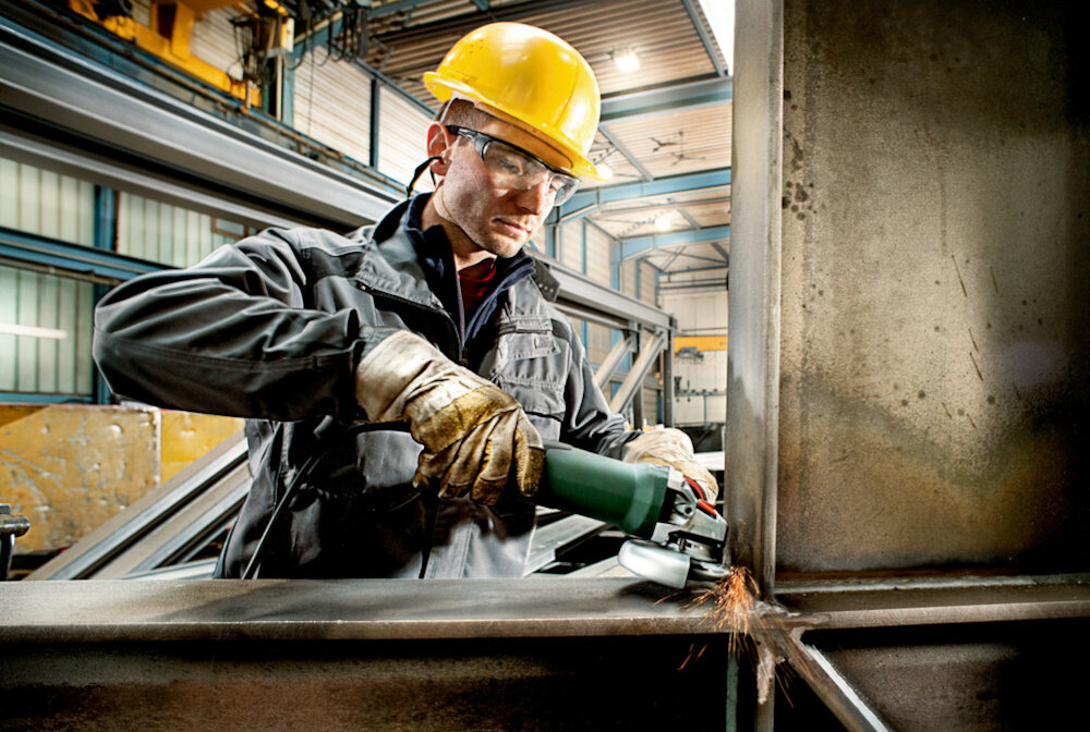 Szlifierka kątowa METABO WEV 15-125 Quick 600468000 osoba trzyma szlifierkę dłoniach tnie metalowy element w tle hala magazyn do cięcia szlifowania wygładzania do pracy w stali i betonie bezpieczne użytkowanie prosta obsługa lekka konstrukcja