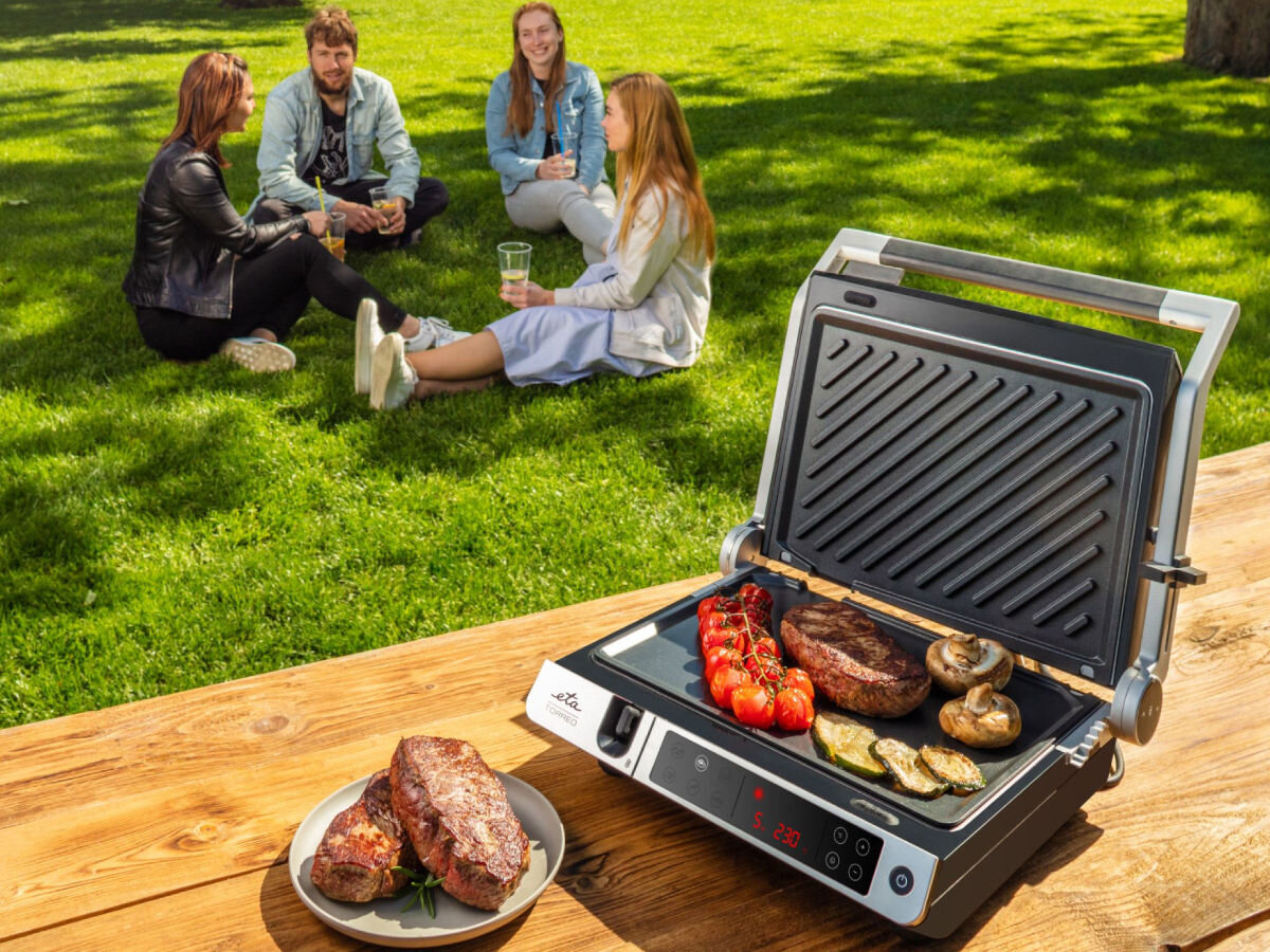 Grill elektryczny ETA Torreo 715590000, kuchnia, gotowanie, ogród, grillowanie, Piknik na trawie – grill ETA Torreo otwarty z jedzeniem na płycie, w tle grupa znajomych, całość w plenerze.