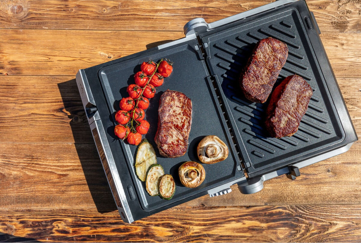 Grill elektryczny ETA Torreo 715590000, regulacja, timer, sonda, wygoda, Widok z góry na całkowicie otwarty grill ETA Torreo 715590000, na płytach steki i warzywa, grill stoi na drewnianym stole.