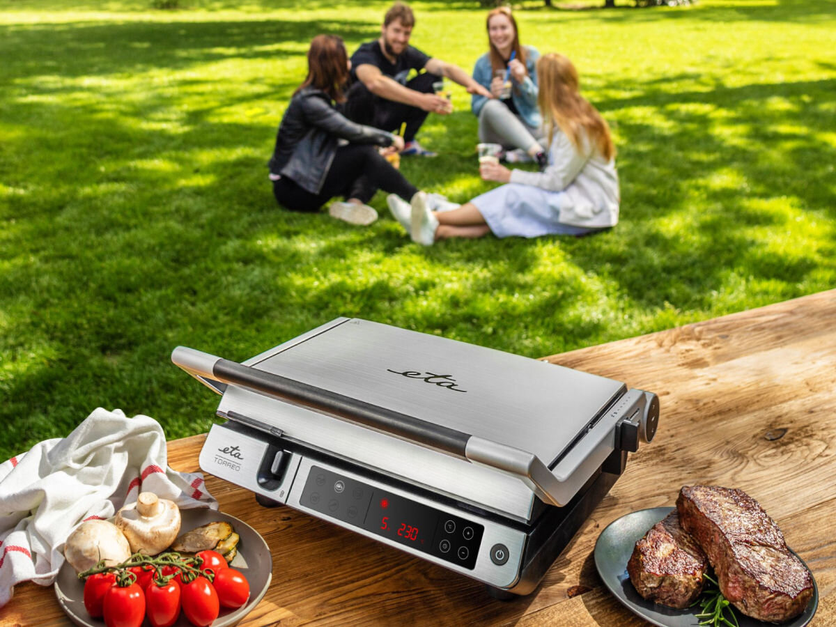 Grill elektryczny ETA Torreo 715590000, bezpieczeństwo, uchwyty, łatwość, mycie, Grill ETA Torreo 715590000 zamknięty, przygotowane składniki i steki na talerzu, w tle cztery osoby na trawie.