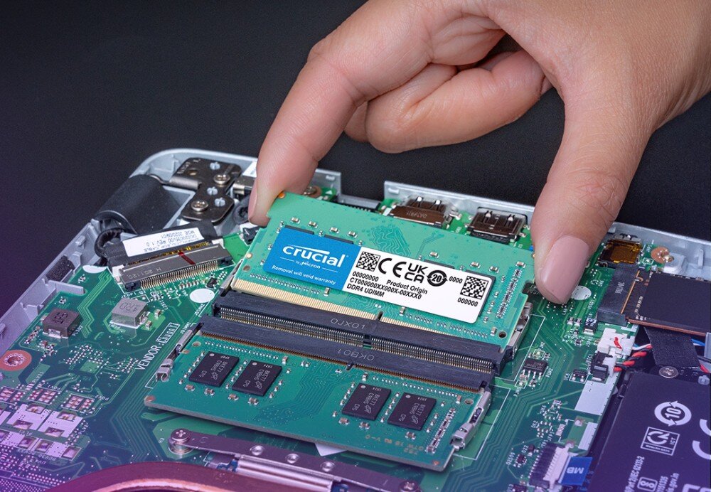 Pamięć RAM CRUCIAL Zbliżenie na dłoń wkładającą zielony moduł pamięci do gniazda w laptopie, widoczne są elementy płyty głównej i złącza, pojemność, gniazda
