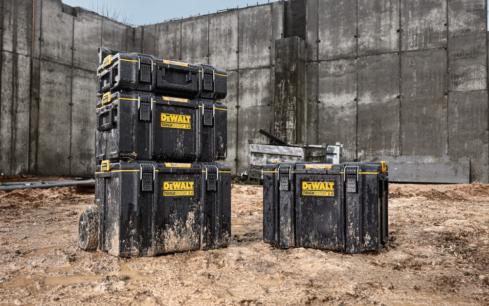 Skrzynka narzędziowa DEWALT DWST83294-1 plac budowy piasek skrzynia ustawiona na piasku obok 3 inne skrzynie ustawione jedna na drugiej należy do systemu ToughSystem 2.0 Toolbox w pełni kompatybilna z produktami ToughSystem pierwszej generacji można łączyć nowe elementy z już posiadanymi modułami dowolnie rozbudowywać zestaw