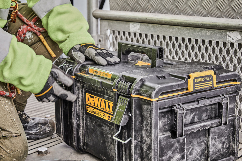 Skrzynka narzędziowa DEWALT DWST83294-1 plac budowy skrzynka ustawiona na ziemi osoba trzyma zamek w dłoni dwuczęściowe solidne metalowe zamki bezpieczne przechowywanie narzędzi i akcesoriów ochrona przed przypadkowym otwarciem podczas przenoszenia trwałość całej skrzynki