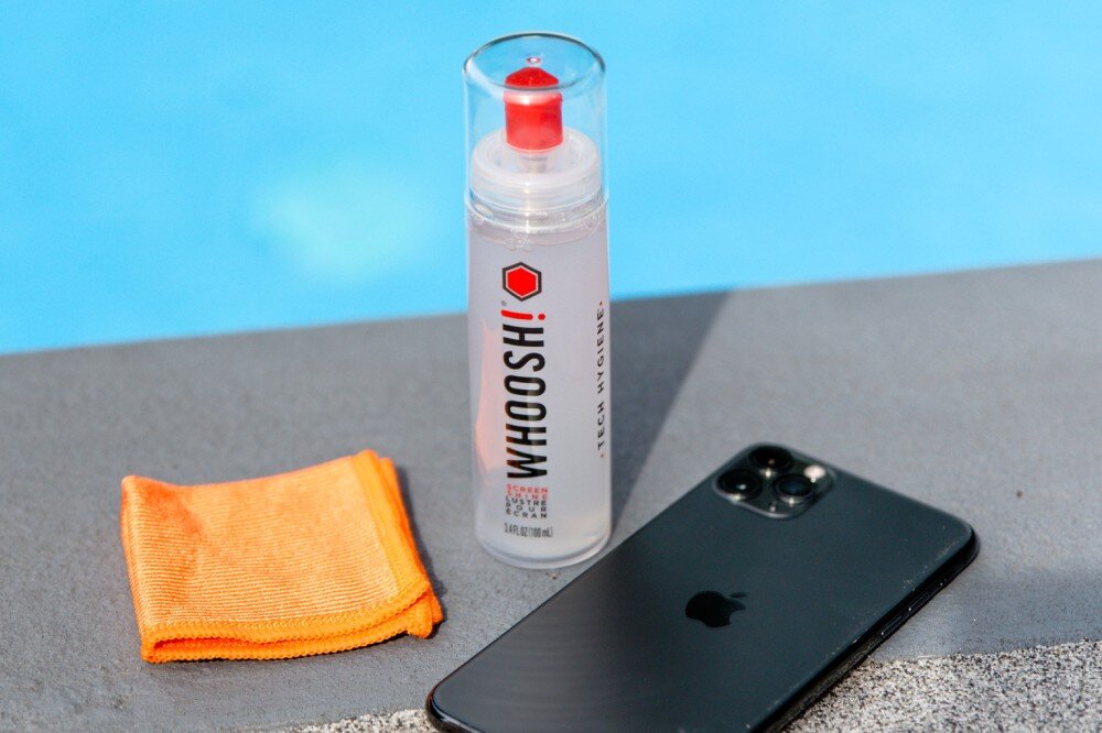 Zestaw czyszczący WHOOSH! do ekranów LCD/TFT/LED Go XL 100 ml Plastikowa butelka z płynem czyszczącym stoi obok pomarańczowej ściereczki oraz ciemnego smartfona na szarym murku przy błękitnej tafli wody