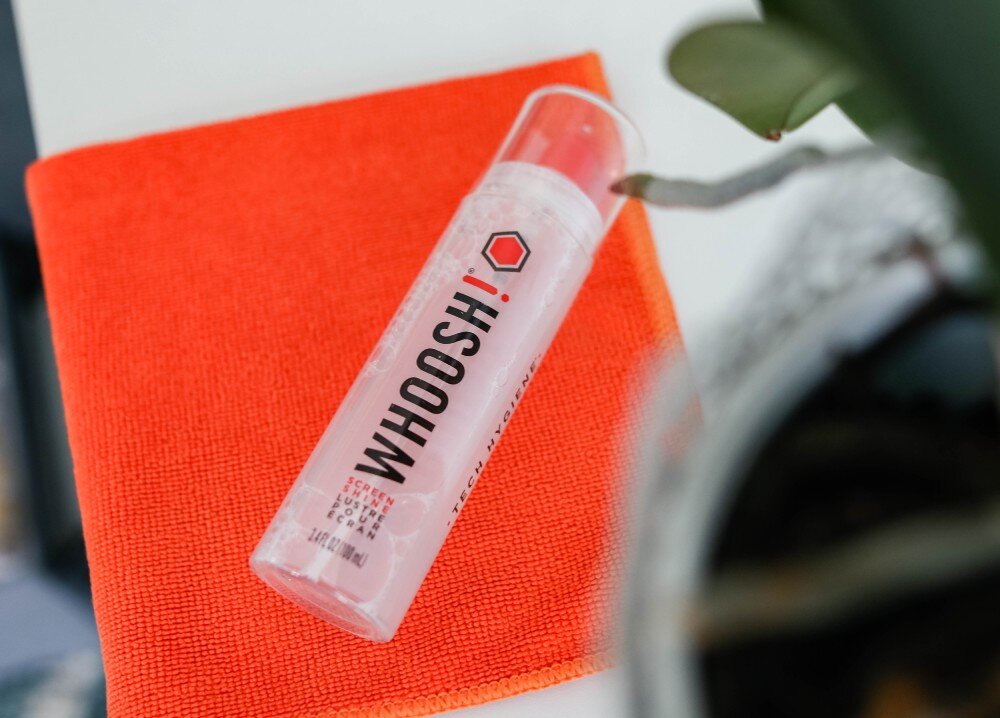 Zestaw czyszczący WHOOSH! do ekranów LCD/TFT/LED Go XL 100 ml Widok z góry na butelkę z płynem czyszczącym położoną na pomarańczowej ściereczce obok doniczki z rośliną