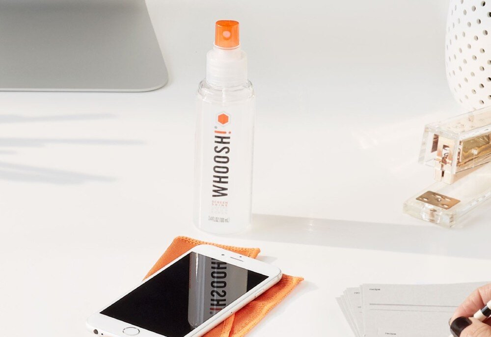 Zestaw czyszczący WHOOSH! do ekranów LCD/TFT/LED Go XL 100 ml Butelka z płynem czyszczącym stoi na białym biurku za smartfonem leżącym na pomarańczowej ściereczce, w otoczeniu biurowych akcesoriów