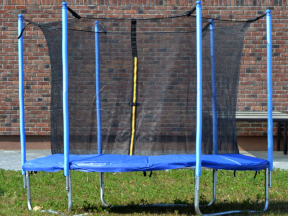 Trampolina ENERO FI 305 cm, skoki, zabawa, aktywność, Trampolina stoi w pełno rozstawiona, na trawniku, w słoneczny dzień, w tle ściana budynku
