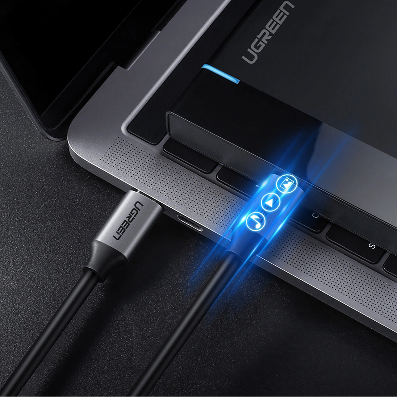 Na obrazie widoczny jest szary kabel USB-C podłączony do laptopa, z logo \'UGREEN\' na wtyczce. Kabel posiada podświetlane ikony na końcówce, a na laptopie widoczne jest logo \'UGREEN\'.