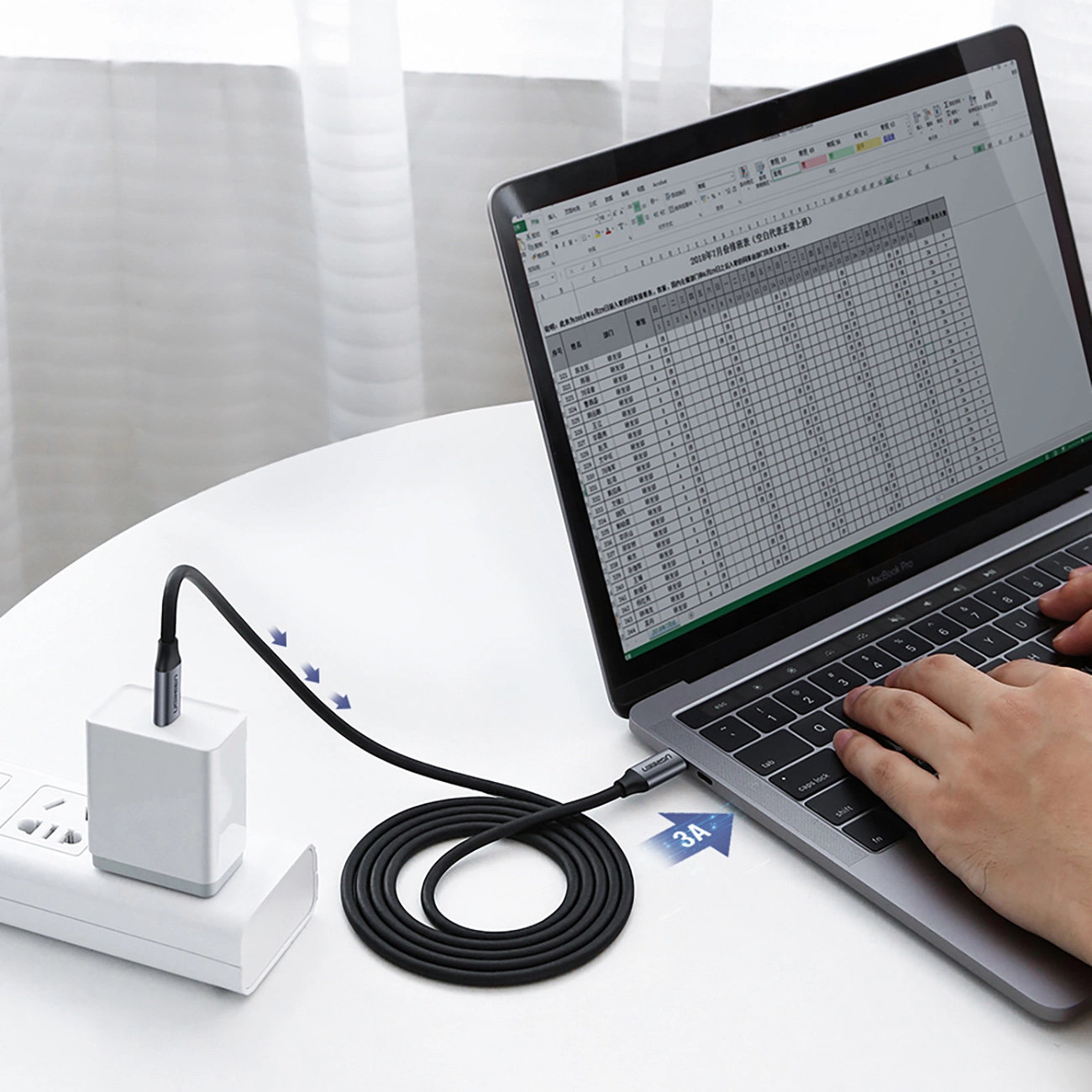 Na białym stole znajduje się laptop z otwartym arkuszem kalkulacyjnym, do którego podłączony jest szary kabel USB-C. Kabel jest również podłączony do białej ładowarki wpiętej w listwę zasilającą.