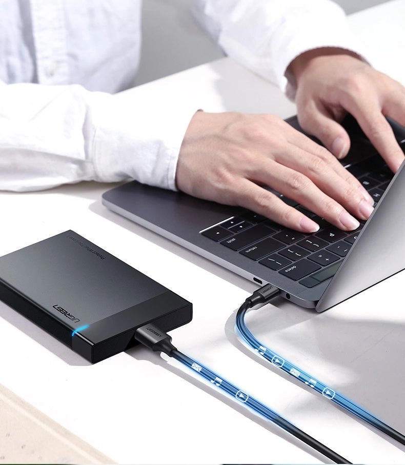 Osoba pisze na laptopie, do którego podłączony jest zewnętrzny dysk twardy za pomocą kabla USB. Na dysku widoczne jest logo \'Ugreen\'.