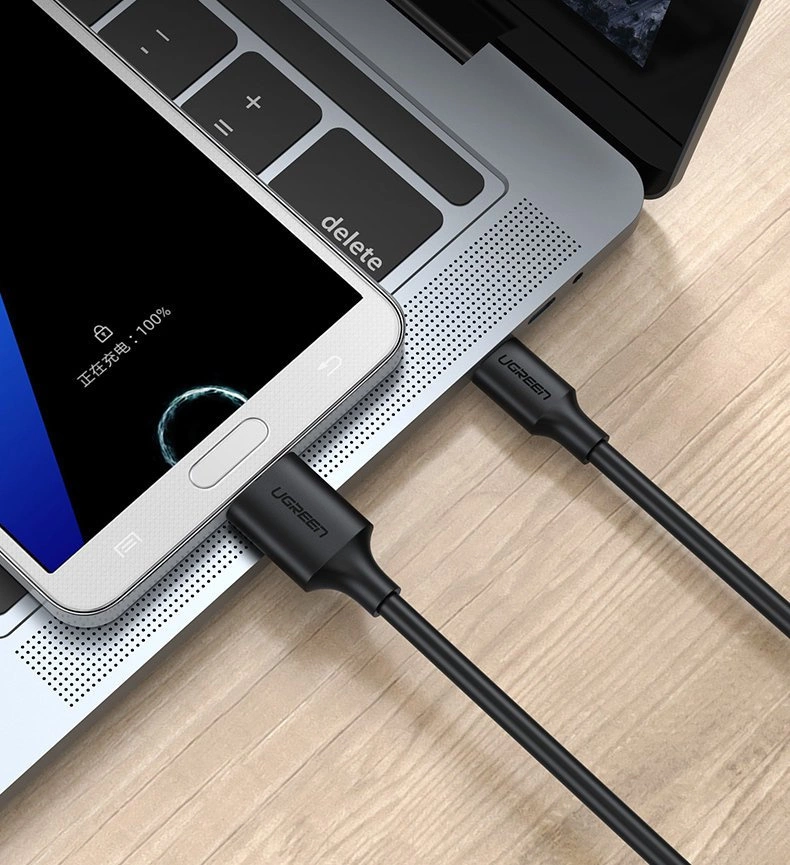 Na obrazie widać czarny kabel USB marki Ugreen podłączony do laptopa i smartfona. Na ekranie smartfona widoczny jest tekst: \'正在充电: 100%\'.