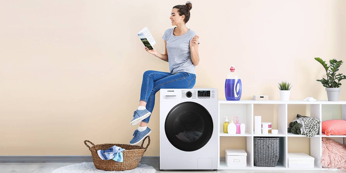 Pralko-suszarka z widocznym logo Samsung stoi obok białego regału z różnymi przedmiotami, w tym butelką detergentu i rośliną. Na pralce siedzi osoba trzymająca ulotkę, a przed nią znajduje się kosz z praniem.