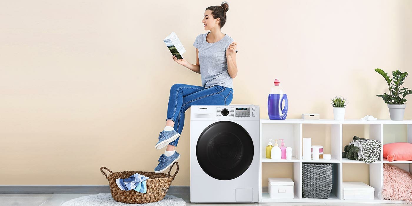 Kobieta siedzi na pralce i czyta książkę, obok na pułkach leżą ręczniki i płyny do mycia 