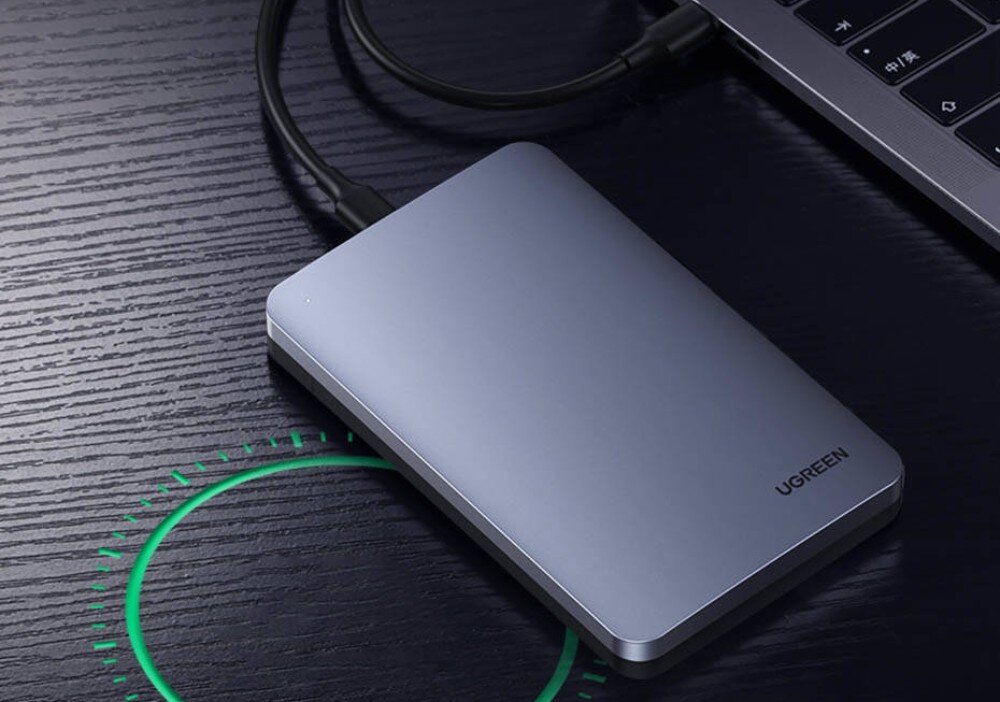 Obudowa dysku UGREEN CM300 70499 Srebrna obudowa dysku zewnętrznego marki Ugreen leżąca na ciemnym blacie obok laptopa i podłączona do niego czarnym kablem