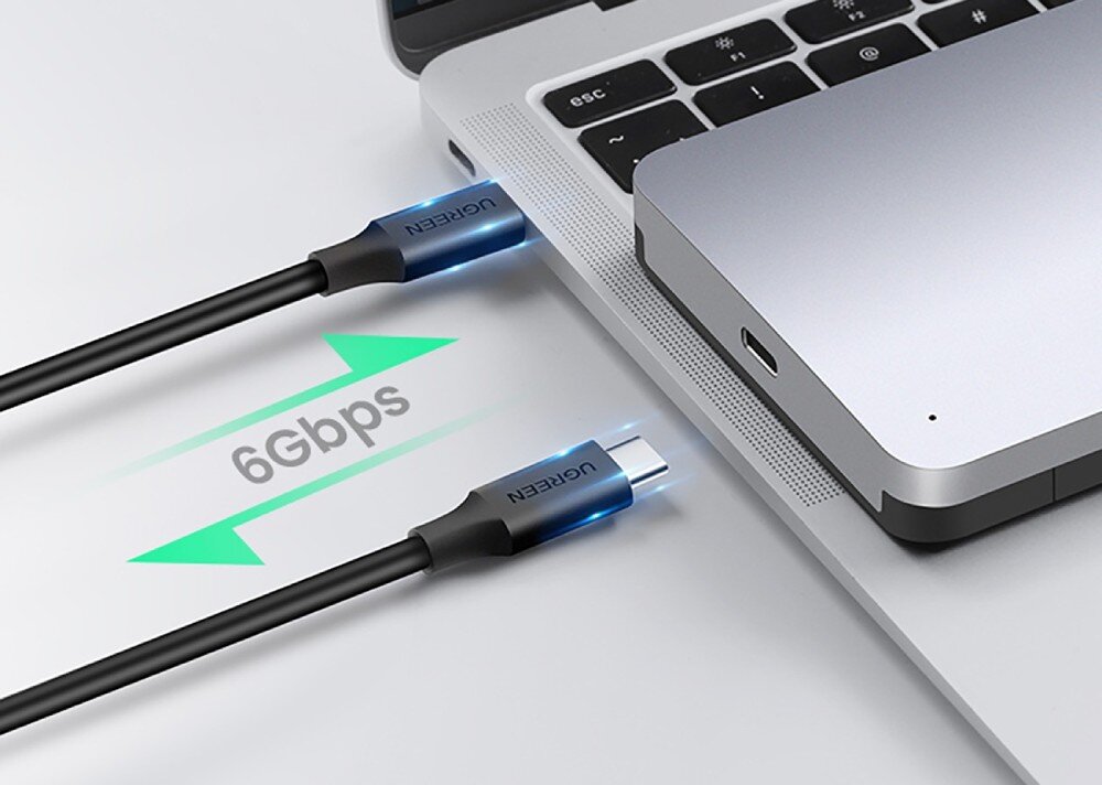Obudowa dysku UGREEN CM300 70499 Zbliżenie na dłoń podłączającą czarny kabel USB-C do laptopa obok którego leży srebrna obudowa dysku a między nimi widnieje zielona ikona strzałek z napisem '6Gbps'