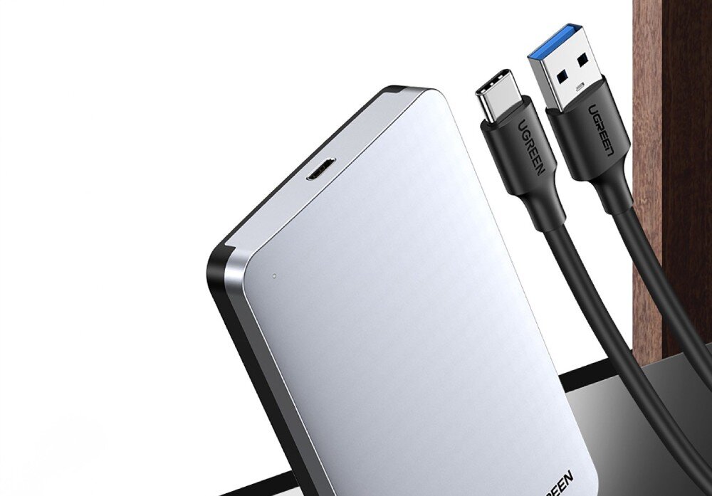 Obudowa dysku UGREEN CM300 70499 Srebrna obudowa dysku zewnętrznego na białym tle obok dwóch czarnych kabli z których jeden posiada końcówkę USB-C a drugi USB-A z niebieskim środkiem