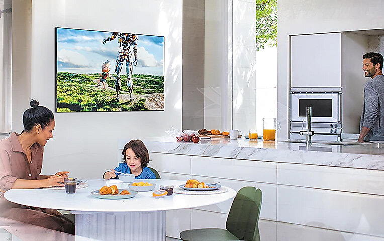 Telewizor wisi na ścianie. Przy stole siedzą matka i dziecko, ojciec stoi obok Q95TDTXXH