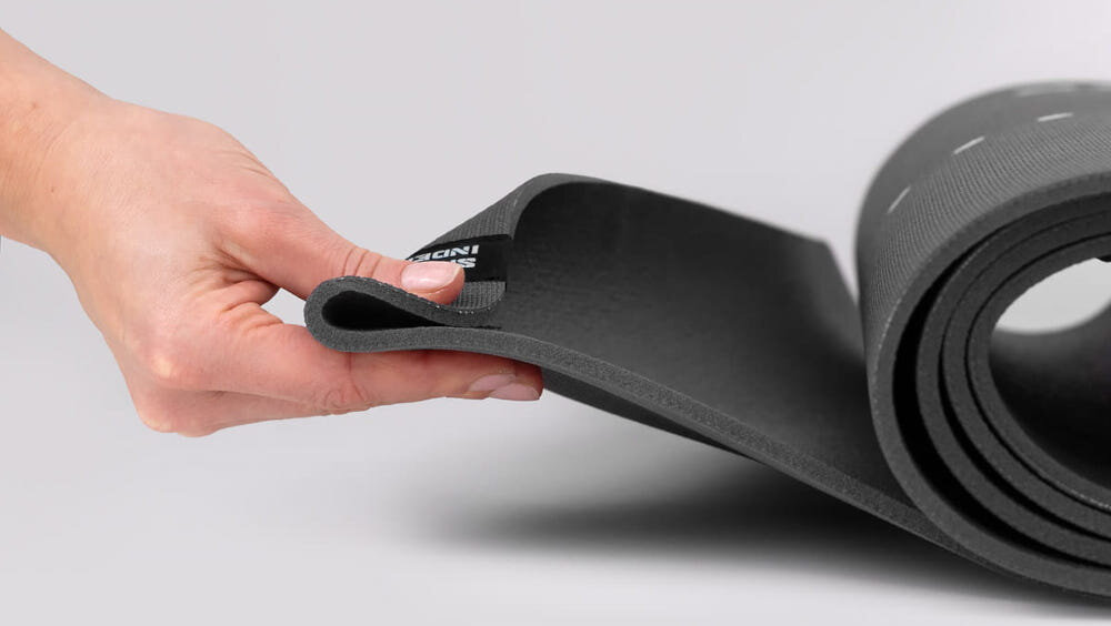 Mata do jogi TIGUAR Yoga Basis Grafitowy jasne tło zwinięta mata osoba trzyma jeden koniec maty w palcach widok z boku wymiary 183 x 60 x 0,5 cm nie ślizga się po podłożu pewność ruchów stabilne oparcie dłoni i stóp zwiększa bezpieczeństwo płynniejszy trening i mniejsze ryzyko niekontrolowanych poślizgów