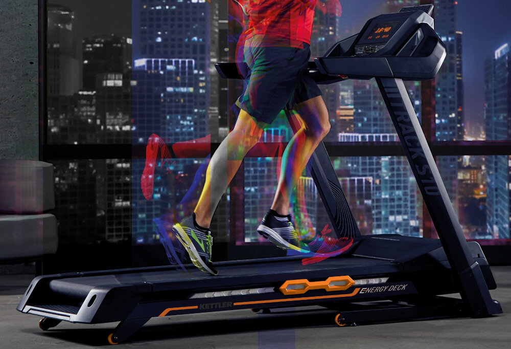 Osoba biegnie na bieżni elektrycznej w nowoczesnym salonie z drewnianą podłogą i dużymi oknami. W tle widoczne są meble, telewizor i lampy.