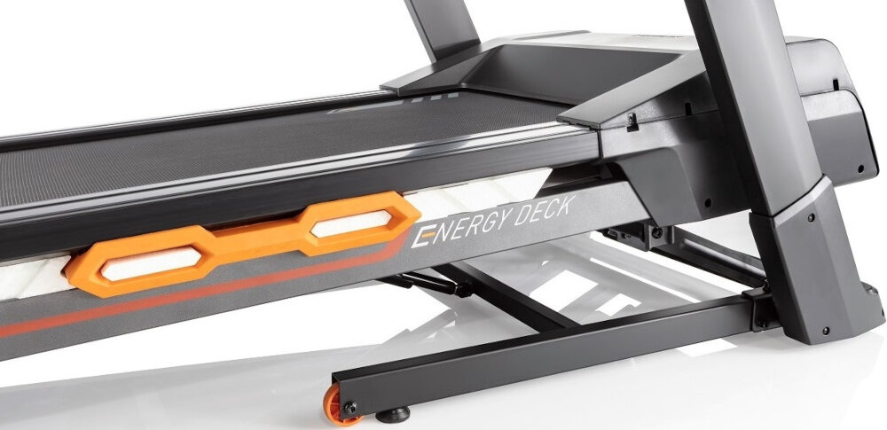 Osoba biegnie na bieżni elektrycznej z widocznym napisem \'ENERGY DECK\' i \'KETTLER\'. W tle znajduje się telewizor i zasłony.