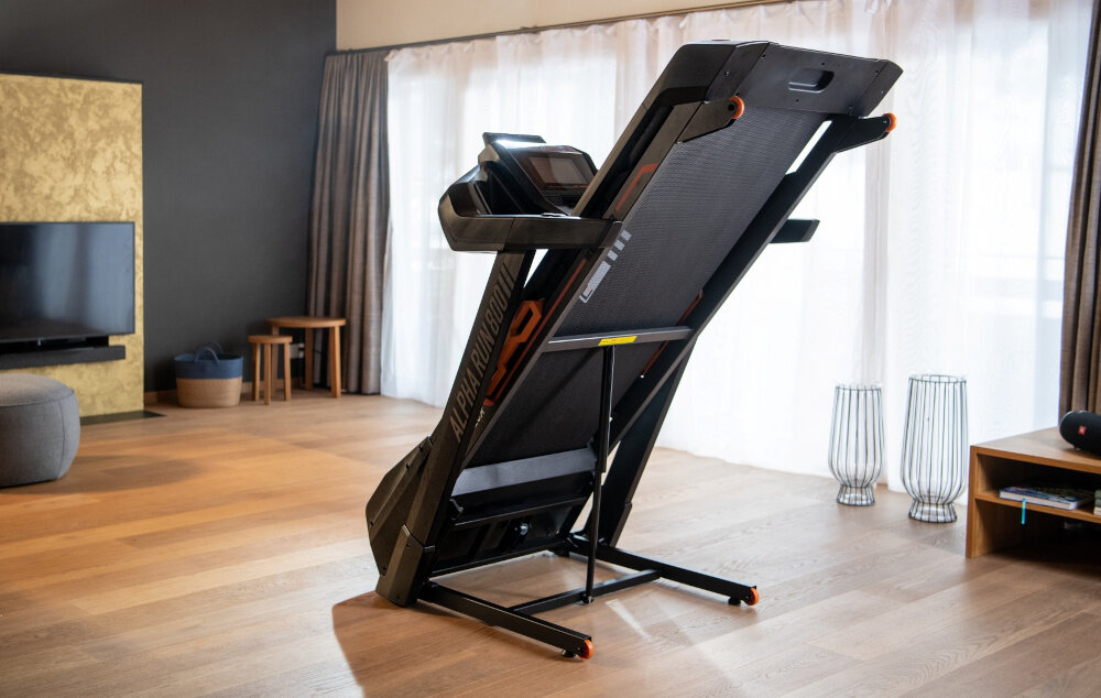 Kobieta stoi przy złożonej bieżni elektrycznej z napisem \'ALPHA RUN 800\' w jasnym, nowoczesnym salonie. W tle widoczne są meble, telewizor i duże okna z zasłonami.