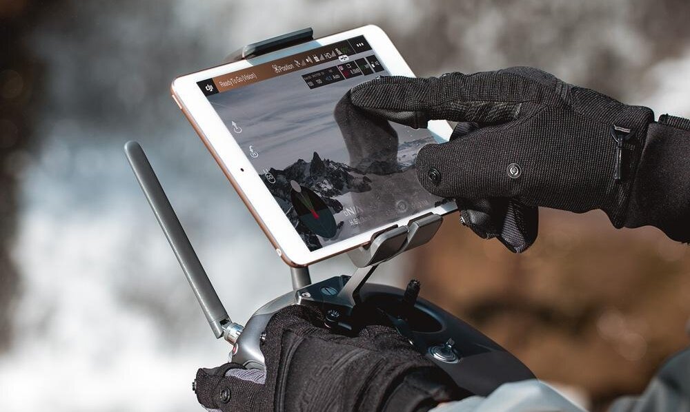 Osoba w czarnych rękawiczkach obsługuje tablet zamocowany na kontrolerze drona. Na ekranie tabletu widoczny jest krajobraz górski oraz interfejs aplikacji do sterowania dronem.