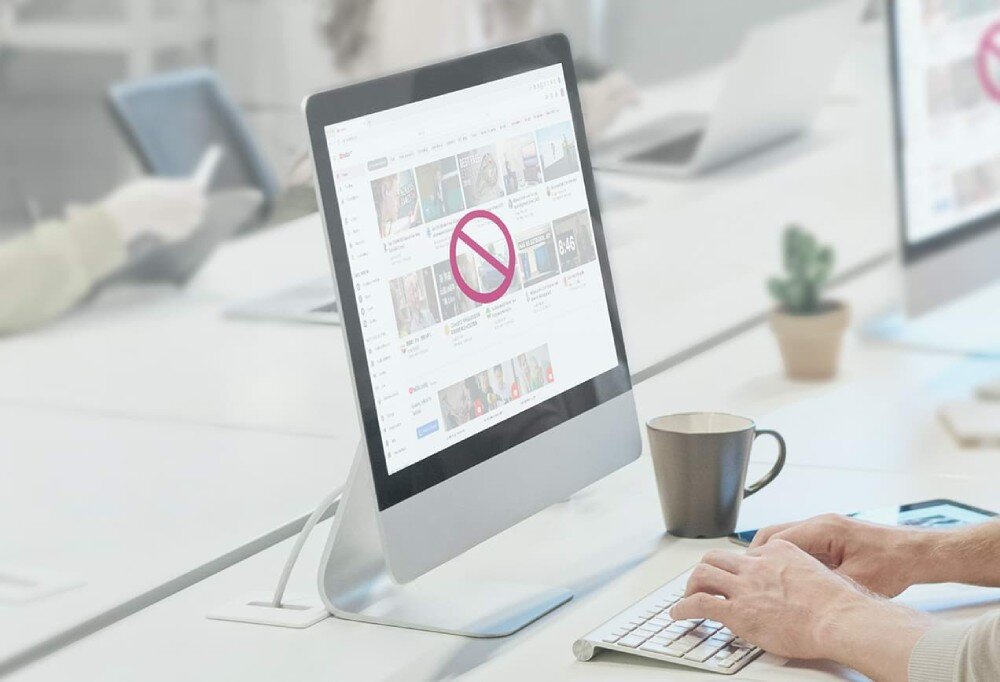 Router TP-LINK TL-ER605 Zdjęcie monitora w biurze z otwartym serwisem wideo. Na ekranie widoczny jest symbol zakazu, sugerujący blokadę strony. W tle pracownicy przy biurkach korzystający z komputerów, kontrola nad siecią, sieci VLAN
