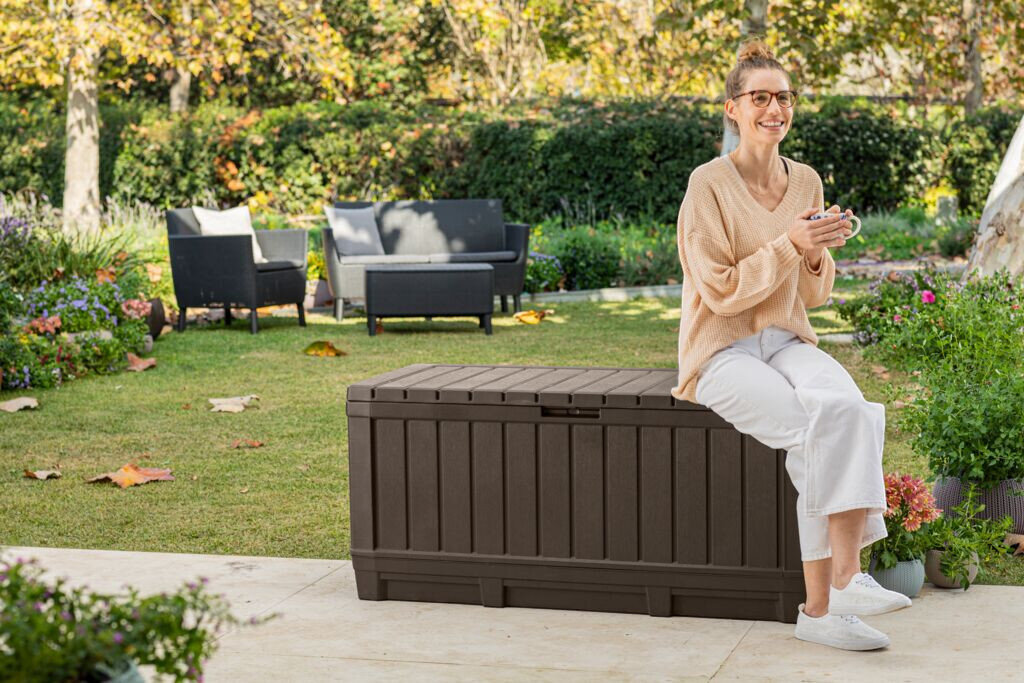 Skrzynia ogrodowa KETER Kentwood 249461 350L Brązowy, siedzisko, obciążenie, ławka, wytrzymałość, Kobieta siedząca na brązowej skrzyni ogrodowej Kentwood w ogrodzie.