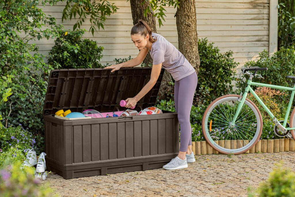 Skrzynia ogrodowa KETER Kentwood 249461 350L Brązowy, kłódka, bezpieczeństwo, uchwyty, udźwig, Kobieta wyjmująca hantle z otwartej skrzyni ogrodowej Kentwood.