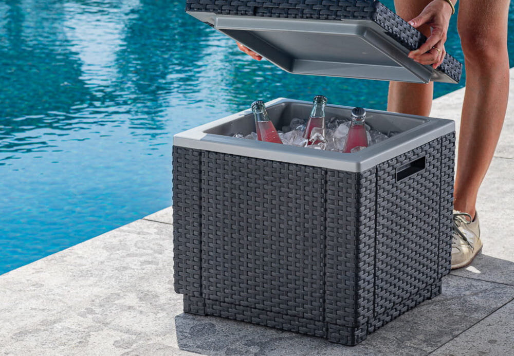 Pufa ogrodowa KETER Ice Cube 213828 Grafitowy pufa z otwartą klapą wnętrze wypełnione kostkami lodu i butelkami w tle basen osoba trzyma klapę w dłoniach praktyczne rozwiązanie wielofunkcyjny mebel trzy funkcje siedzisko lodówka schowek