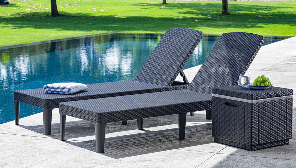 Pufa ogrodowa KETER Ice Cube 213828 Grafitowy pufa z zamkniętą klapą na blacie szklanka z wodą i talerz z winogronami obok dwa leżaki zdjęcie na tle basenu i ogrodu faktura inspirowana rattanem komponuje się z meblami ogrodowymi przyjemna w dotyku łatwa do czyszczenia waga około 3,5 kg uchwyty