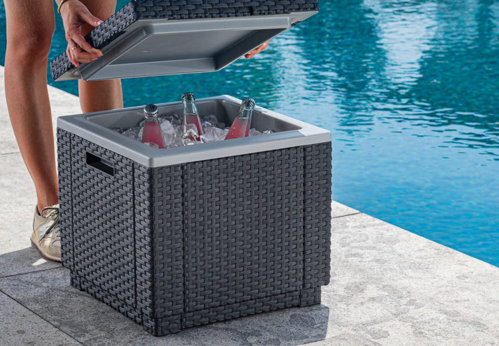 Pufa ogrodowa KETER Ice Cube 213828 Grafitowy pufa z otwartą klapą wnętrze wypełnione kostkami lodu i butelkami w tle basen osoba trzyma klapę w dłoniach z wytrzymałego tworzywa sztucznego nie odbarwia się pod wpływem promieni UV odporne na deszcz i zmiany temperatur na zewnątrz przez cały sezon