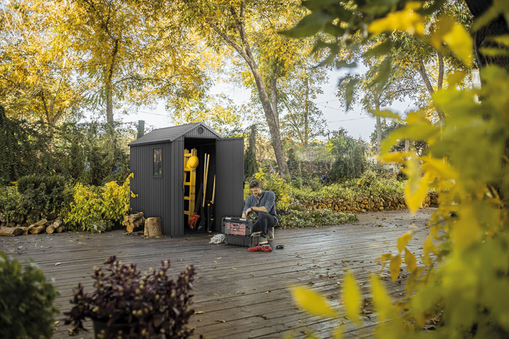 Na drewnianym tarasie stoi szary domek narzędziowy z otwartymi drzwiami, w którym widać narzędzia ogrodowe. W tle znajdują się drzewa z żółtymi liśćmi, a obok domku siedzi osoba z narzędziami.