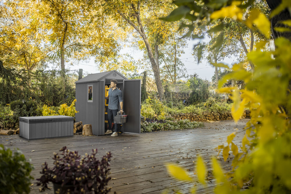 Szary domek narzędziowy stoi na drewnianym tarasie w otoczeniu zieleni i drzew. Obok domku znajduje się mężczyzna trzymający skrzynkę z narzędziami, a w tle widoczne są krzewy i kamienny mur.