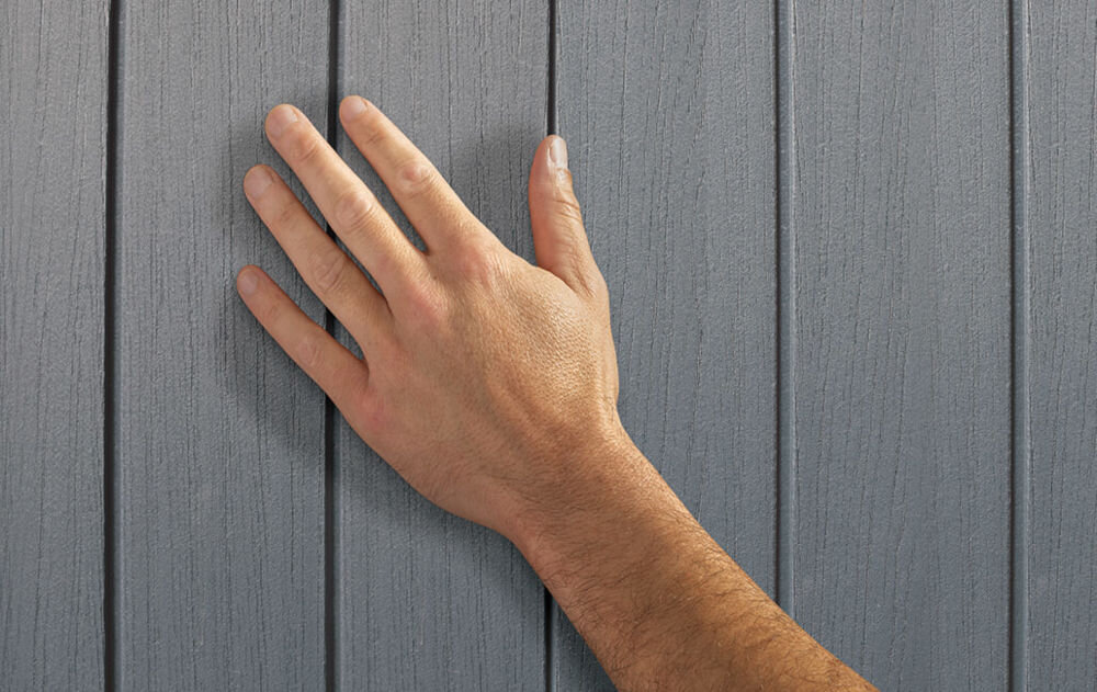 Dłoń dotyka szarej, pionowej powierzchni z teksturą przypominającą drewno. Widoczne są wyraźne linie oddzielające poszczególne panele.