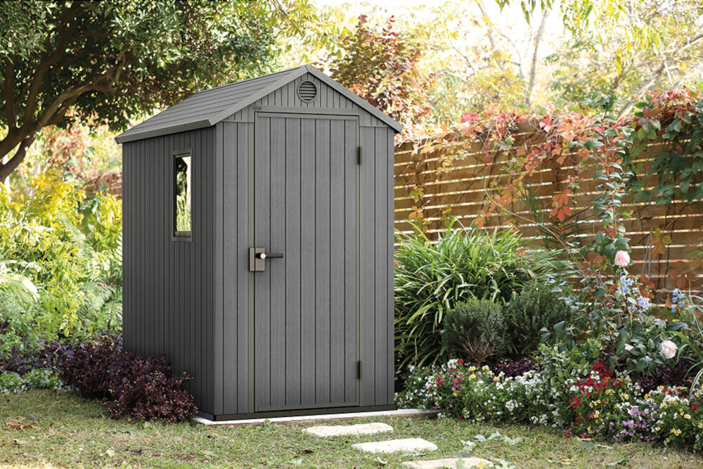 Szary domek narzędziowy z oknem i drzwiami, otoczony bujną roślinnością i kwiatami. W tle widoczny jest drewniany płot.